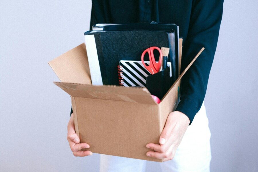 Woman carrying cardboard box with office items like scissors and notebooks.