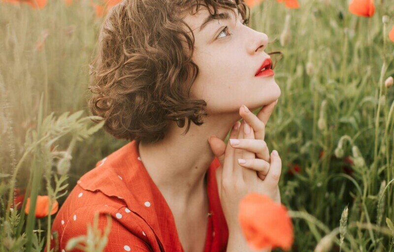 Contemplative woman in a poppy field, wearing a red polka dot dress, surrounded by vibrant flowers.