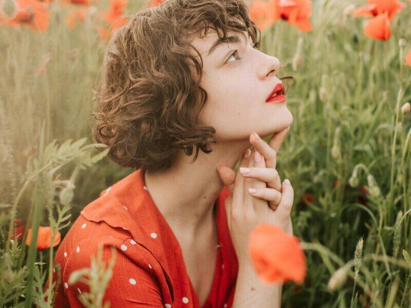 Contemplative woman in a poppy field, wearing a red polka dot dress, surrounded by vibrant flowers.