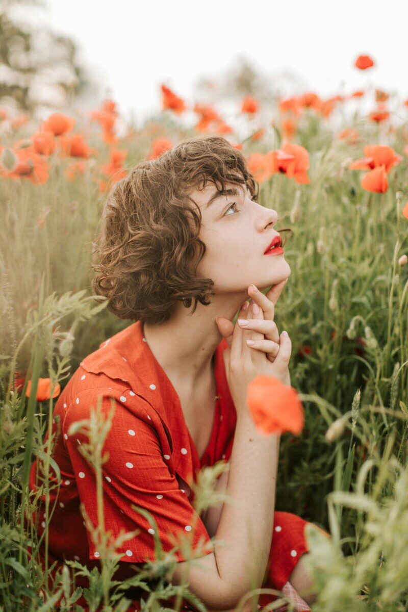 Contemplative woman in a poppy field, wearing a red polka dot dress, surrounded by vibrant flowers.