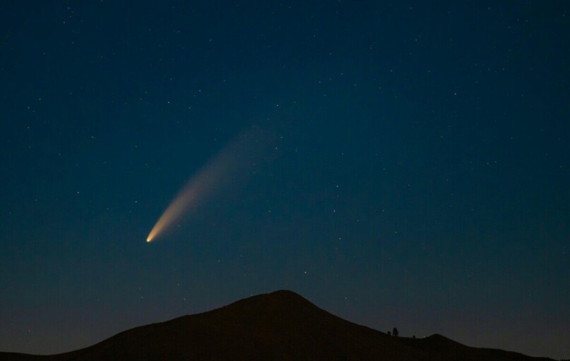 A breathtaking capture of a comet streaking across a star-filled night sky above a mountain silhouette.