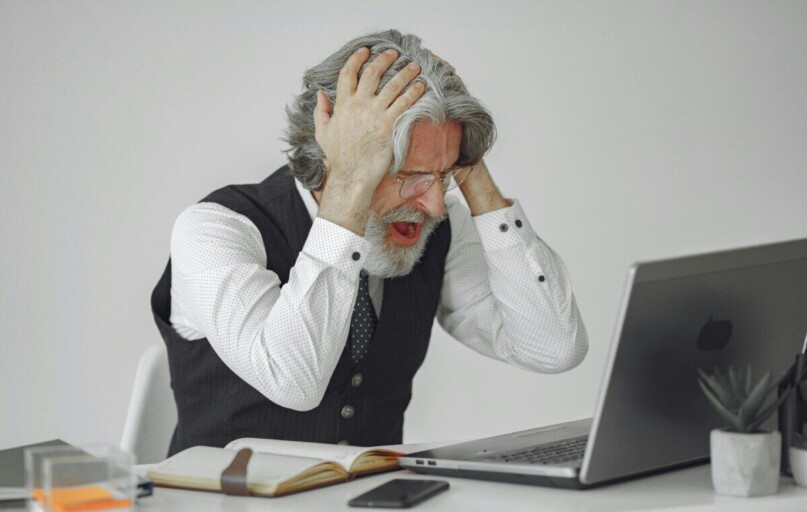 Senior businessman sitting at desk, expressing frustration with his laptop in a professional office setting.