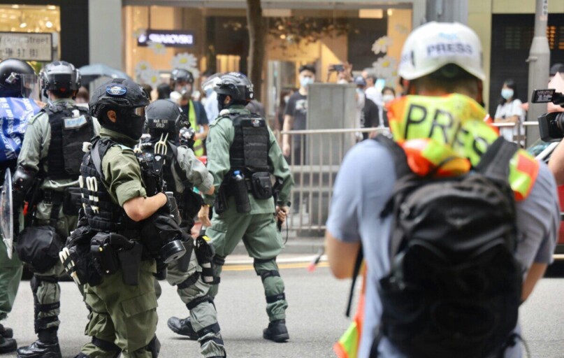 Riot police and journalists during a city protest, wearing protective gear.