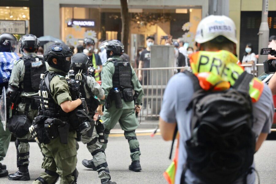 Riot police and journalists during a city protest, wearing protective gear.