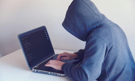 Side view of unrecognizable hacker in hoodie sitting at white table and working remotely on netbook in light room near wall