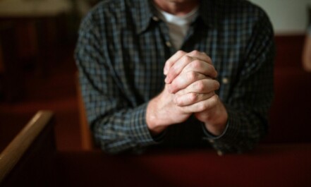 A man in a checked shirt praying in a church, hands clasped together.