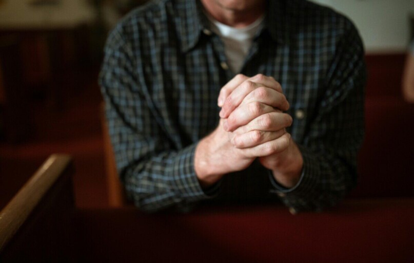 A man in a checked shirt praying in a church, hands clasped together.