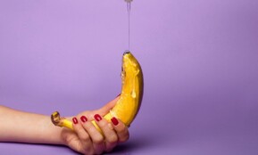 Vivid photo of a hand holding a banana with syrup pouring over it against a purple background.