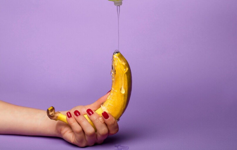 Vivid photo of a hand holding a banana with syrup pouring over it against a purple background.