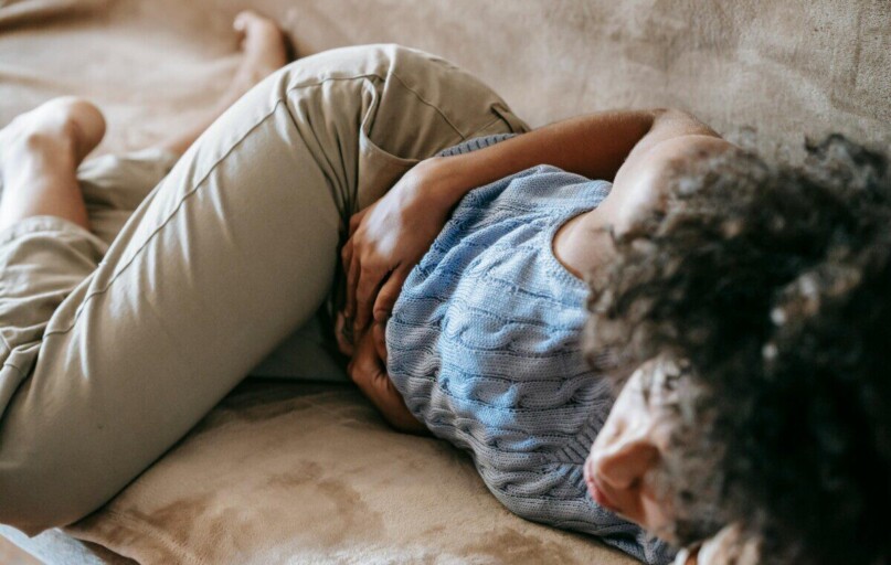 From above of crop anonymous African American female lying on couch and touching belly while having stomach ache