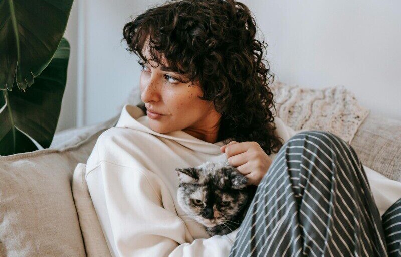 Adult contemplative female embracing cute cat while sitting on soft couch and looking away in living room