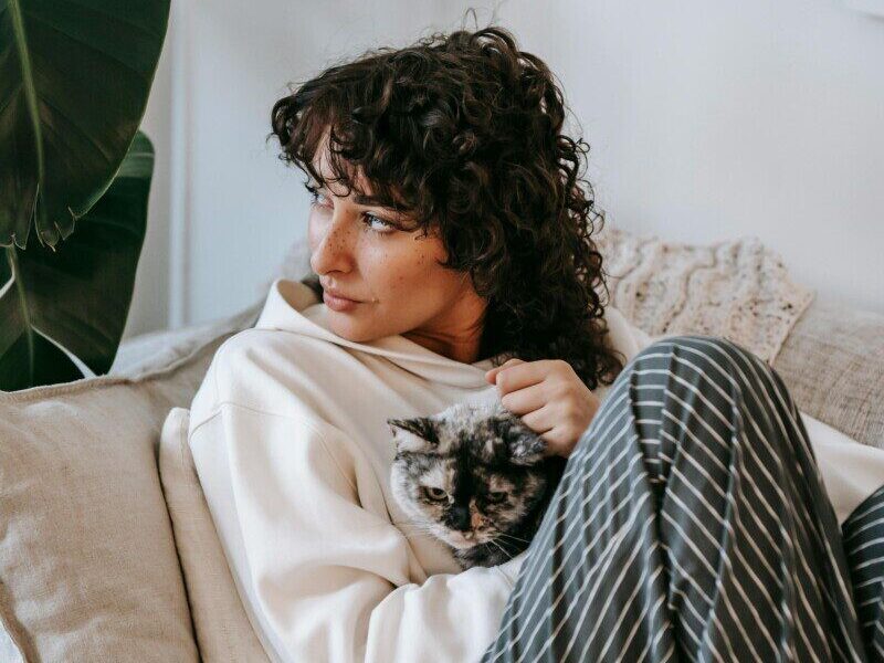 Adult contemplative female embracing cute cat while sitting on soft couch and looking away in living room