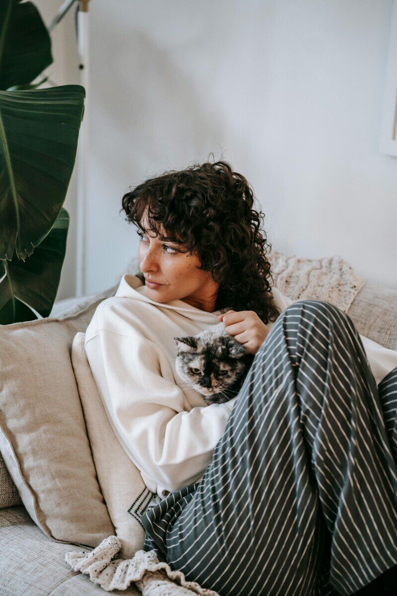 Adult contemplative female embracing cute cat while sitting on soft couch and looking away in living room