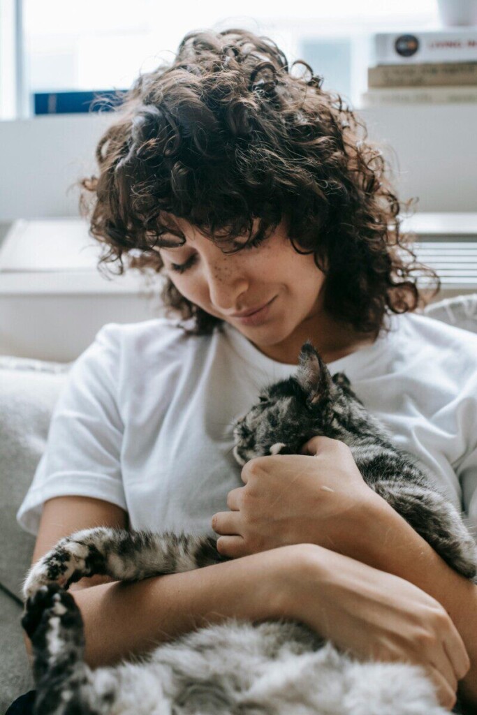 A woman sitting indoors, smiling as she cuddles her calm and content pet cat.