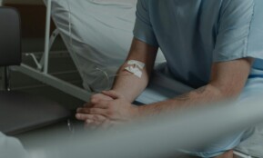 An unrecognizable man in a hospital gown sits on a bed with an IV drip in his arm.