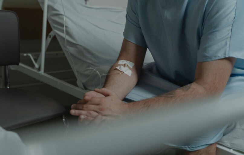 An unrecognizable man in a hospital gown sits on a bed with an IV drip in his arm.