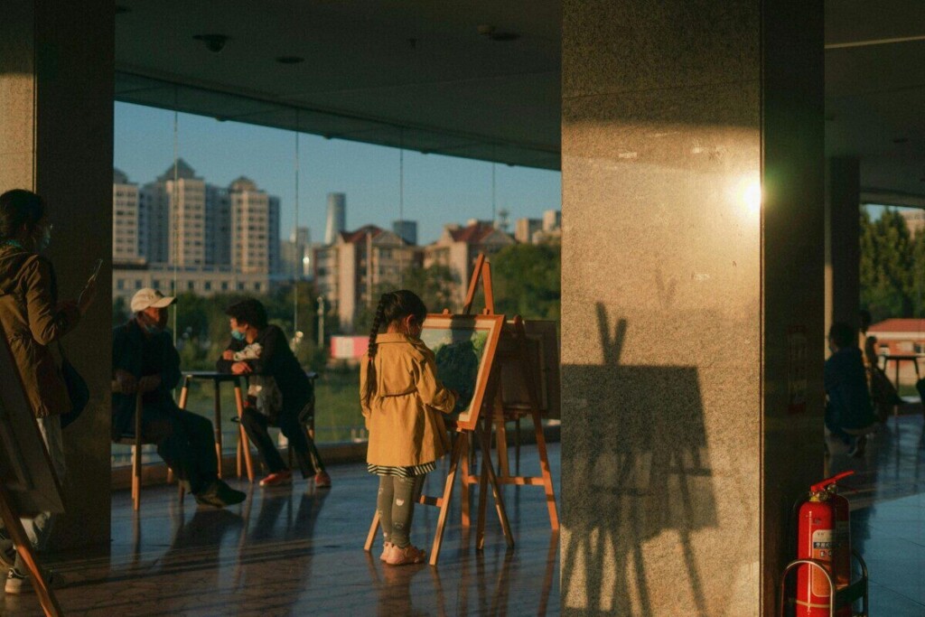 A young girl paints in a sunlit museum space surrounded by art enthusiasts.