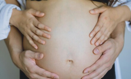 Unrecognizable pregnant wife in black underwear and anonymous gentle husband touching bare belly with hands while standing in room near wall