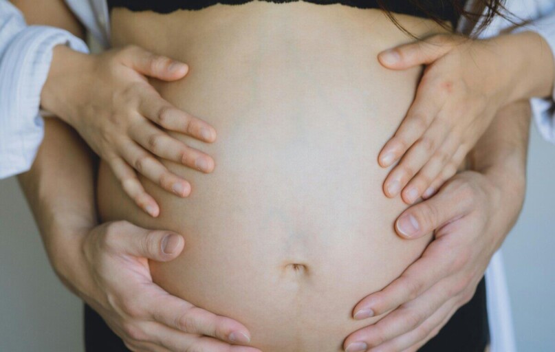 Unrecognizable pregnant wife in black underwear and anonymous gentle husband touching bare belly with hands while standing in room near wall