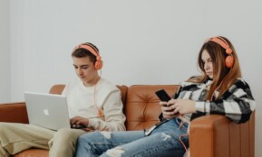 Teens using headphones with laptop and smartphone, lounging on a sofa.