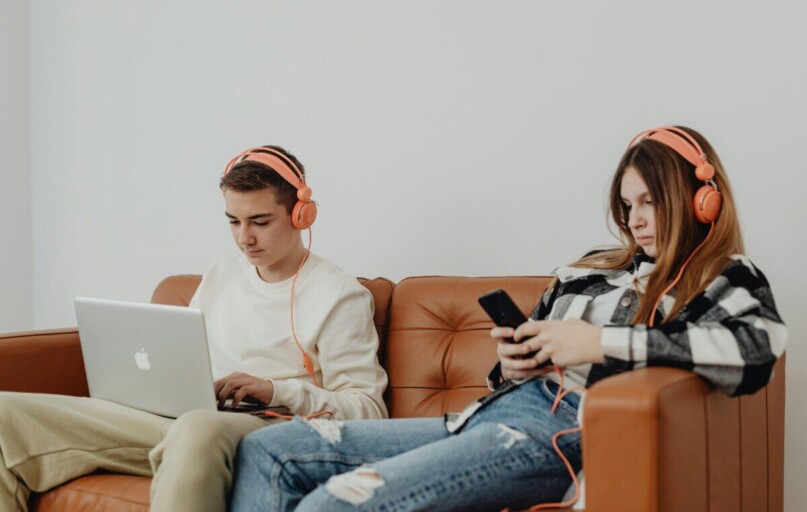 Teens using headphones with laptop and smartphone, lounging on a sofa.