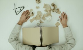 Overhead view of an exhausted man resting with a notepad over his head and crumpled papers around.