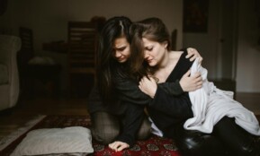 Two women sit closely together indoors, offering comfort and support to each other.