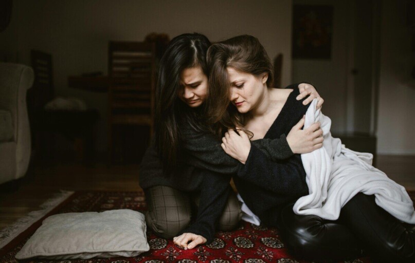 Two women sit closely together indoors, offering comfort and support to each other.