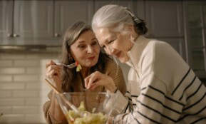 Two senior women smiling and enjoying a fresh salad in a cozy kitchen setting.
