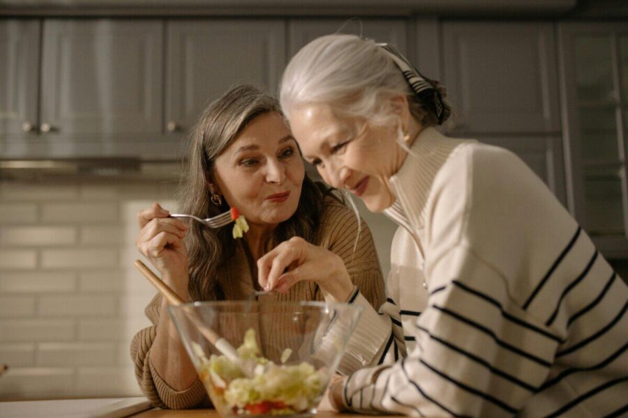 Two senior women smiling and enjoying a fresh salad in a cozy kitchen setting.