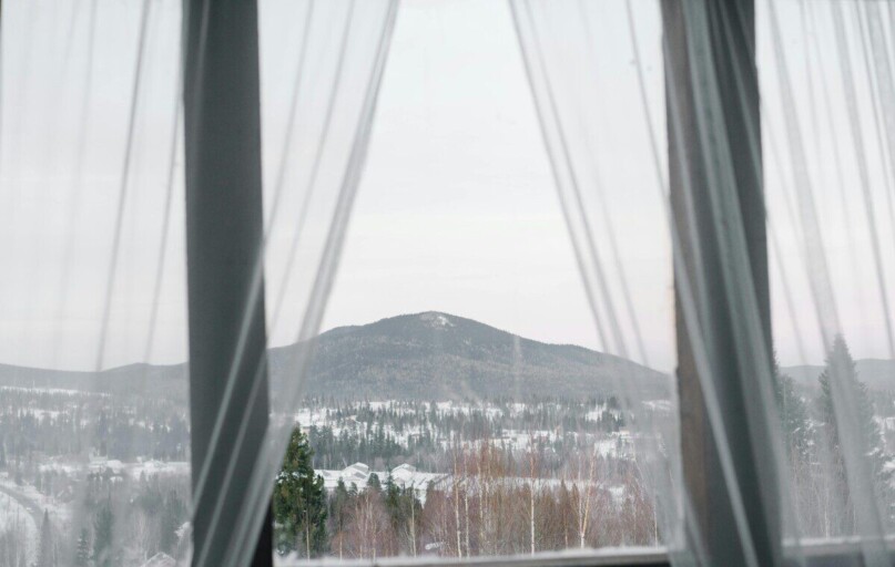 A serene winter landscape framed by sheer curtains, showcasing snow-covered mountains and forests.
