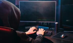 A person in a hoodie sits at a computer screen, engaged in coding or hacking activities.