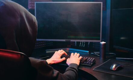 A person in a hoodie sits at a computer screen, engaged in coding or hacking activities.