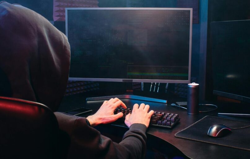 A person in a hoodie sits at a computer screen, engaged in coding or hacking activities.