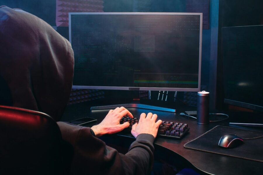 A person in a hoodie sits at a computer screen, engaged in coding or hacking activities.