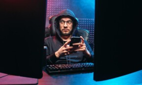 A man in a hoodie using a smartphone, surrounded by tech gear in a dimly lit room.