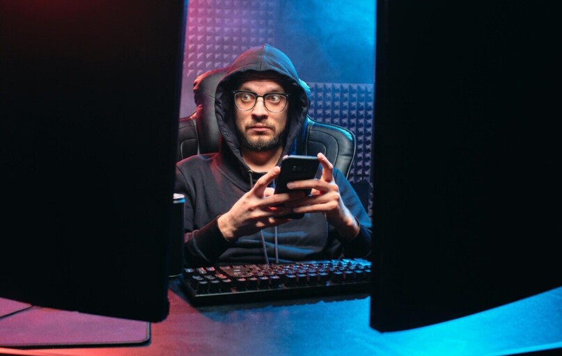 A man in a hoodie using a smartphone, surrounded by tech gear in a dimly lit room.