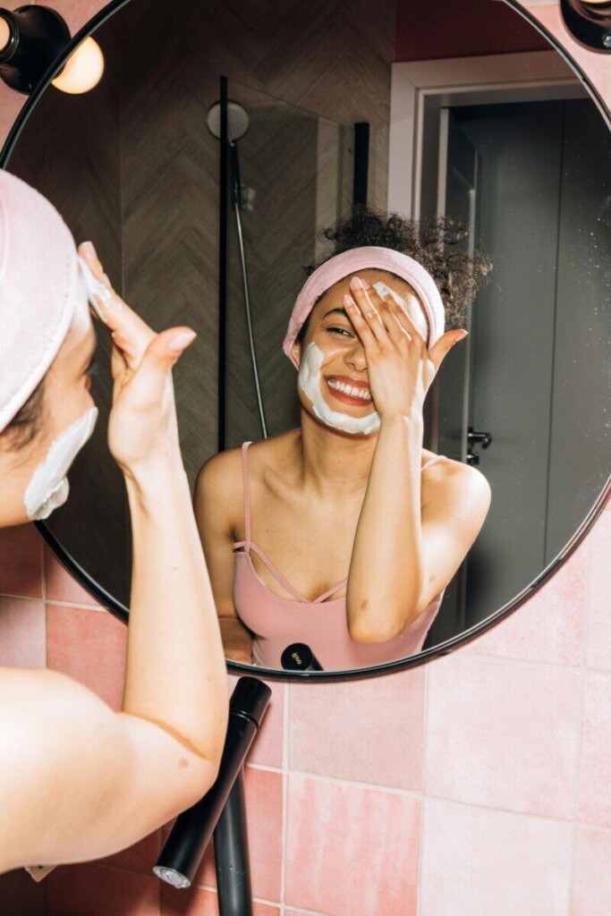 Happy woman applying face mask while smiling in bathroom mirror. Perfect for skincare and beauty concepts.