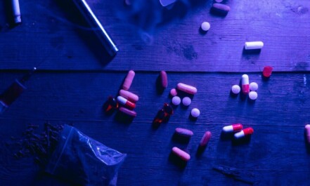A flat lay of various drugs including pills, capsules, and a syringe on a wooden surface under moody blue lighting.