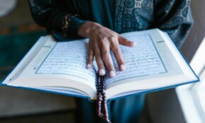 A woman's hand rests on an open Quran with prayer beads, symbolizing devotion.