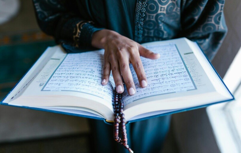 A woman's hand rests on an open Quran with prayer beads, symbolizing devotion.