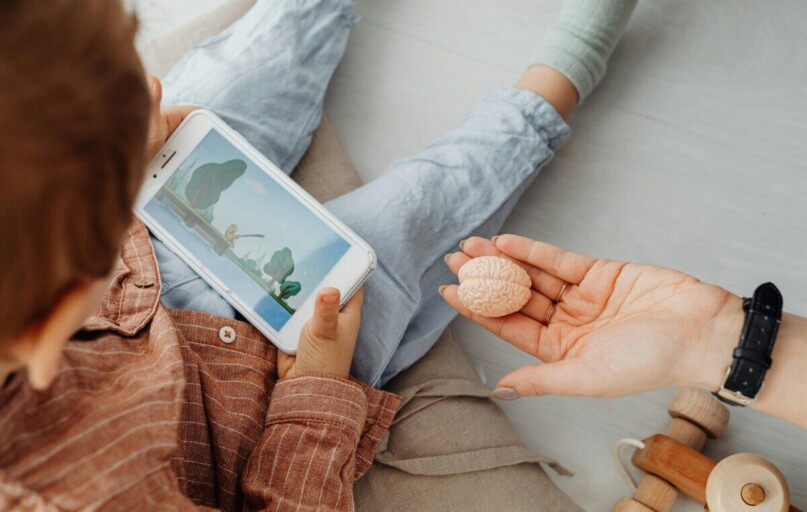 Young child engrossed in a smartphone video while holding a brain model indoors.