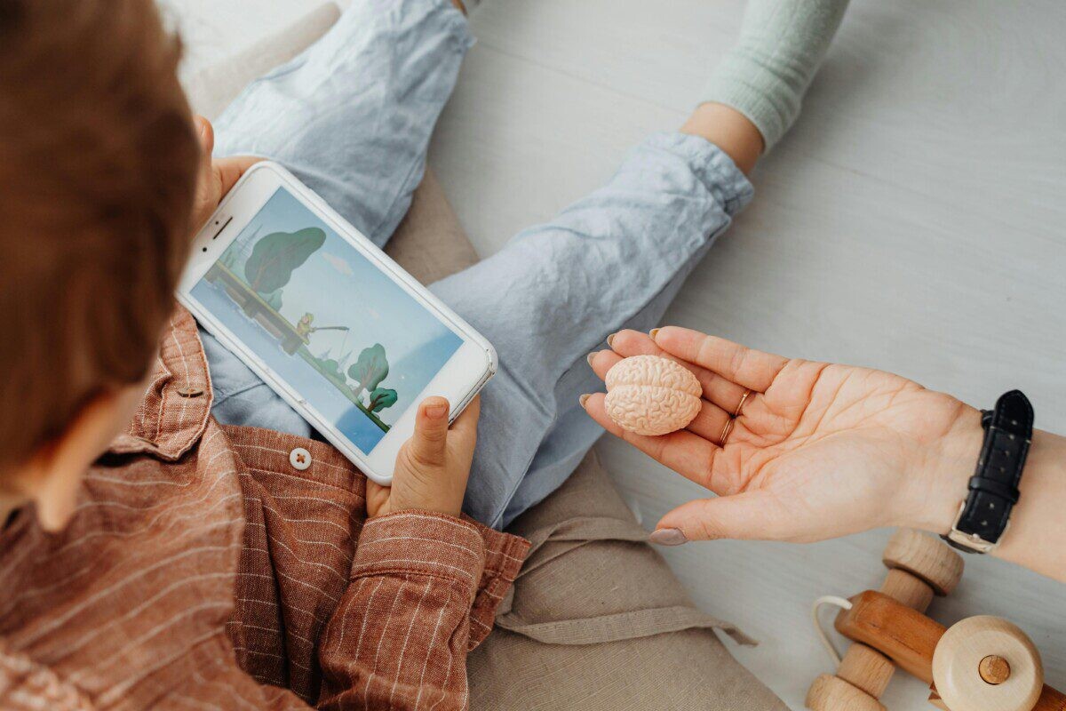 Young child engrossed in a smartphone video while holding a brain model indoors.