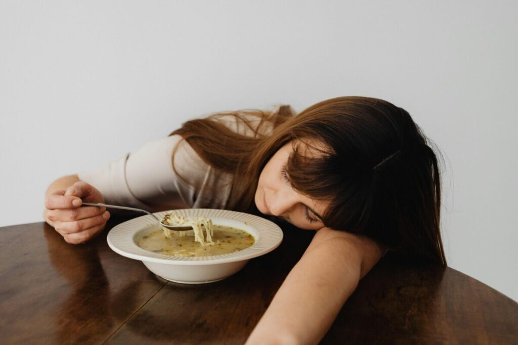 A woman resting her head on the table while holding a spoon and eating soup, conveying fatigue.