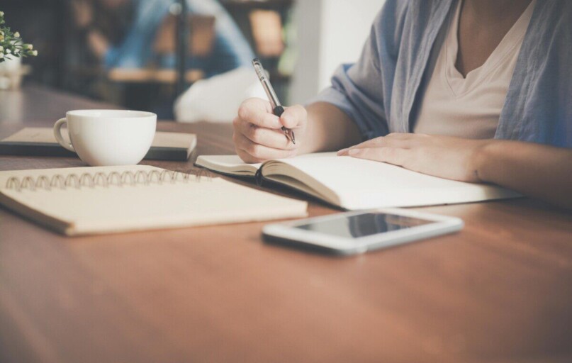 A woman writes in a notebook at a café table with a coffee and smartphone nearby.