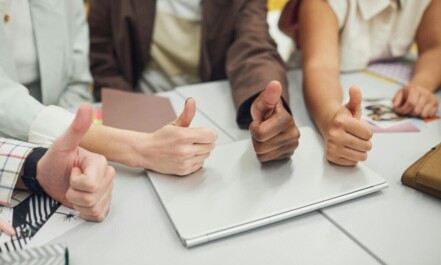 Four diverse hands showing thumbs up over a laptop on a desk, symbolizing teamwork and approval.