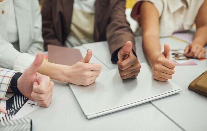 Four diverse hands showing thumbs up over a laptop on a desk, symbolizing teamwork and approval.