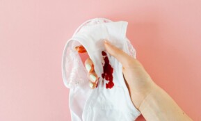 Close-up of a hand holding white underwear with menstrual stain against pink background, highlighting health awareness.