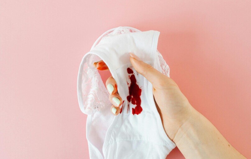 Close-up of a hand holding white underwear with menstrual stain against pink background, highlighting health awareness.