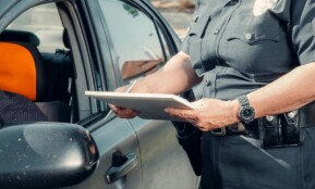A police officer in uniform writes a ticket to a driver through the car window on a sunny day.
